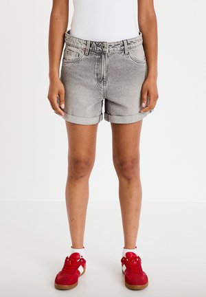 Persoon draagt opgevouwen grijze spijkershorts, wit topje, witte sokken en rode sneakers staand op een witte achtergrond.