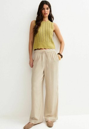 Vrouw met lang donker haar, gekleed in een mouwloos geel gebreid topje, losse beige broek, bruine armbanden en beige platte schoenen tegen een witte achtergrond.