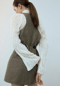 Bruin en beige geruite jurk met een aansluitende silhouet, gelaagd met een doorschijnende witte blouse met getextureerde mouwen en een zwarte riem als accent.