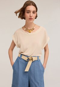 Blusa beige a maniche corte con risvolti abbinata a pantaloni a gamba larga azzurri chiari con una cintura beige e grandi accessori in oro.