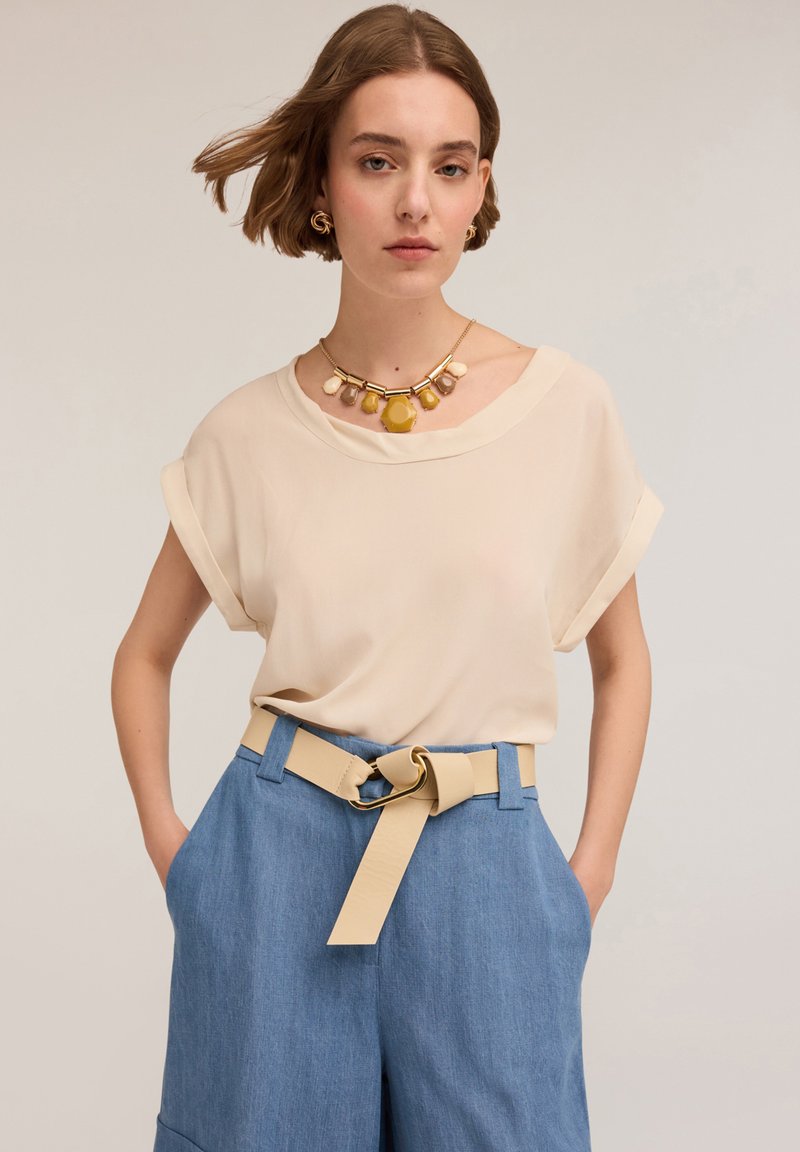 Blusa beige a maniche corte con risvolti abbinata a pantaloni a gamba larga azzurri chiari con una cintura beige e grandi accessori in oro.