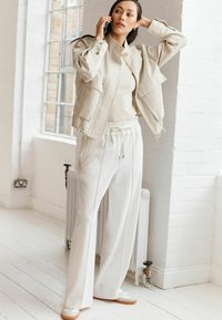 Lichtbeige verkorte jas met twee voorkruiken, gecombineerd met crèmekleurige broek met hoge taille en witte sneakers met beige zolen. Heldere interieursetting.