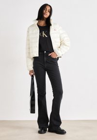 Casaco puffer bege claro com gola alta, combinado com uma t-shirt preta com um logótipo dourado, jeans pretos à boca de sino e sapatos pretos robustos.