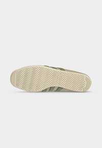 Semelle de sneaker beige avec motif à chevrons, logo Adidas visible au centre et bandes vertes partiellement visibles sur le côté.
