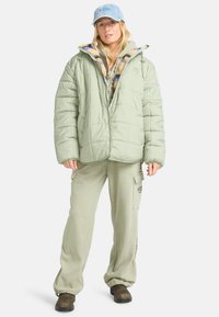 Lichtgroen gewatteerd pufferjack met een capuchon, gelaagd over een gestreept shirt, gecombineerd met bijpassende broek en bruine schoenen. Blauwe pet als accent.