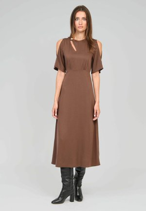 Femme portant une robe midi marron avec des découpes d'épaules dénudées et un petit orifice en forme de clé, associée à des bottes à talons noires montant jusqu'aux genoux, debout sur un fond gris.