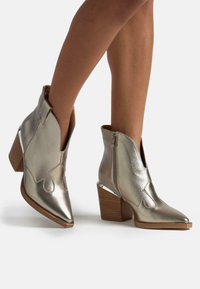 Botas de tobillo plateadas metálicas con punta afilada, costuras decorativas, cierre con cremallera y un tacón de madera apilada con un acento transparente.