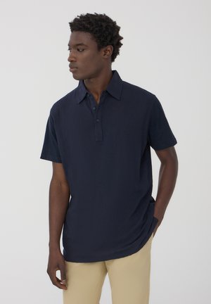 Polo shirt blu navy a maniche corte con tessuto texturizzato, abbottonatura e colletto classico. Abbinato a pantaloni beige.