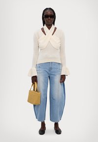 Maglione crema con scollatura drappeggiata, maniche a campana; jeans in denim blu chiaro a vestibilità larga; borsa intrecciata gialla; ballerine nere.