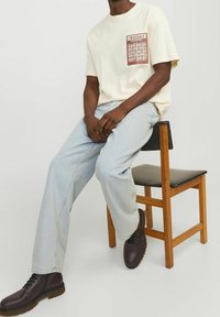 Camiseta crema con un diseño gráfico de bolsillo marrón, combinada con jeans azul claro y botas de cuero marrón oscuro con cordones, sentada en una silla de madera.