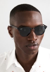 Occhiali da sole neri Ray-Ban con lenti angolari, montatura nera sottile e dettaglio del logo in argento, indossati da una persona con capelli corti e un orecchino.