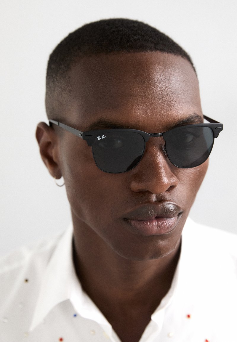 Occhiali da sole neri Ray-Ban con lenti angolari, montatura nera sottile e dettaglio del logo in argento, indossati da una persona con capelli corti e un orecchino.