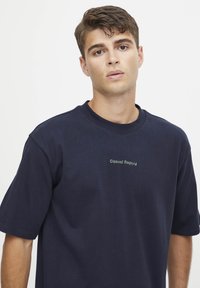 Marineblå bomulls t-skjorte med kortermet og rund hals. Har subtil "Casual Friday"-tekst trykket i grønt på forsiden.