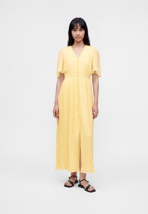 Femme aux longs cheveux foncés portant une robe midi plissée jaune à manches courtes et des sandales noires, debout sur un fond uni.