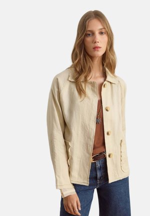 Vrouw met lang lichtbruin haar die een beige jack met knopen, een bruin overhemd, blauwe spijkerbroek en een ketting met een sterrenhanger draagt.