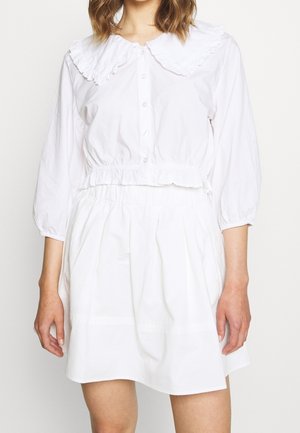 Mujer con blusa blanca abotonada con un gran cuello con volantes y mangas abullonadas combinada con una falda blanca de talle alto.