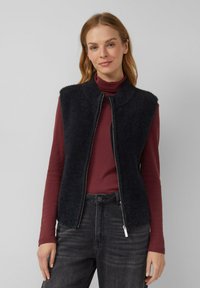 Schwarze, ärmellose Weste aus einem strukturierten, flauschigen Material mit hohem Kragen, ausgestattet mit einem Reißverschluss vorne. Getragen über einem burgunderfarbenen Langarmshirt.