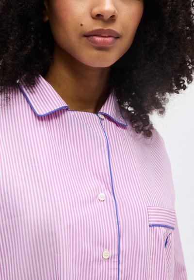 Femme aux cheveux bouclés portant une chemise rayée rose et blanche avec un passepoil bleu et des boutons blancs, montrant le haut du torse et les lèvres.