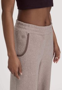 Hellbraune Fleece-Hosen mit hohem Bund, ausgestattet mit einer Seitentasche, die von dunklen braunen Nähten umrandet ist, sowie einem kleinen Logo auf der Tasche.