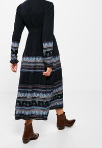 Pitkä, tumma mekko, jossa on kuvioitu helma ja hihat sinisen ja maanläheisten sävyjen yhdistelmänä, istuva yläosa ja levenevä hame. Yhdistetty ruskeisiin nilkkabootseihin.