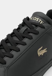 Baskets Lacoste noires avec lacets noirs, texte Lacoste doré sur la languette et logo crocodile doré sur le côté.