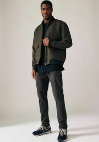Jachetă bomber verde închis cu un model texturat, cămașă lungă neagră dedesubt, blugi gri închis și adidași bleumarin cu accente albe.