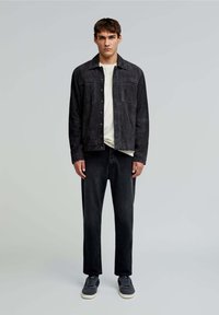 Jachetă din suede negru, cu două buzunare frontale, purtată peste un tricou de culoare deschisă, asociată cu blugi negri și pantofi gri. Fundal simplu.