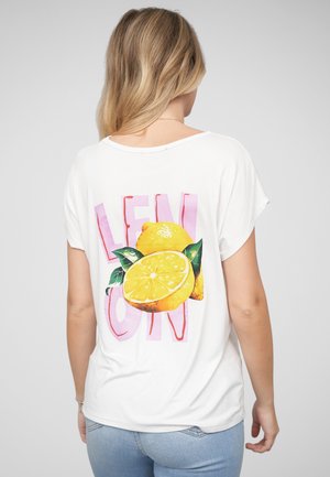 Frau mit blonden Haaren, die ein weißes T-Shirt mit großen gelben Zitronengrafiken und pinkem Text auf dem Rücken trägt, kombiniert mit hellblauen Jeans.