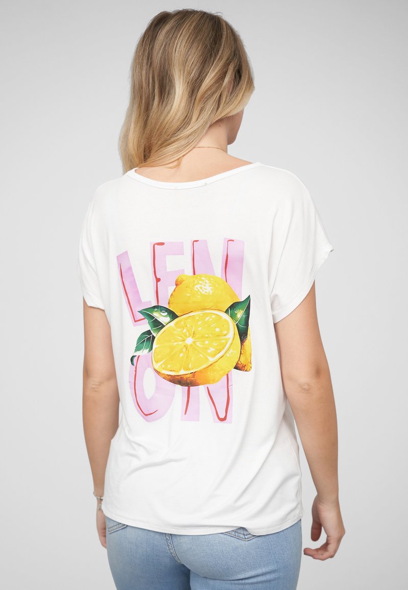 Femme aux cheveux blonds portant un t-shirt blanc avec de grands graphismes jaunes de citron et un texte rose dans le dos, associé à un jean bleu clair.
