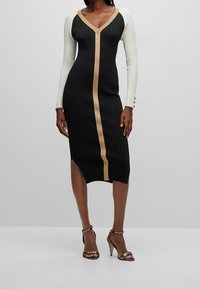 Robe en maille côtelée noire avec manches longues blanches et une bande verticale dorée. Présente un décolleté en V et une fente à l'ourlet.