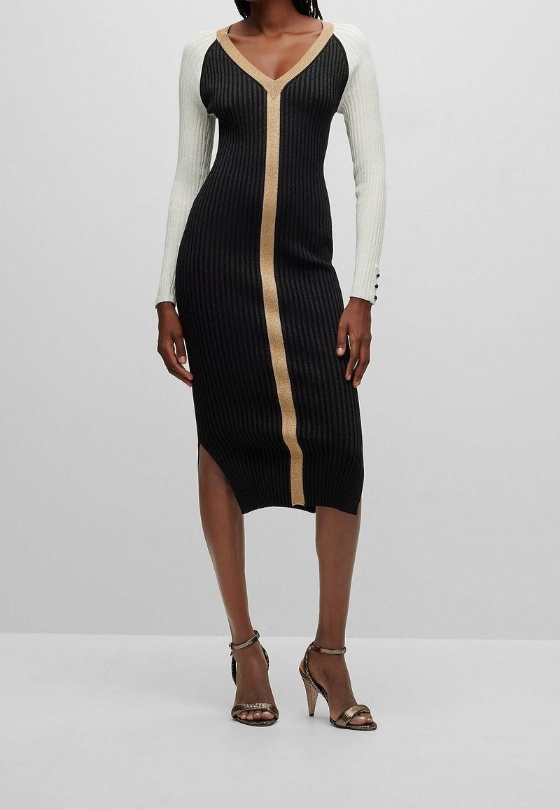 Robe en maille côtelée noire avec manches longues blanches et une bande verticale dorée. Présente un décolleté en V et une fente à l'ourlet.