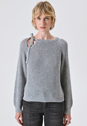 Donna con capelli biondi corti, che indossa un maglione grigio a maglia con dettaglio sulla spalla annodato e jeans scuri, in piedi contro uno sfondo neutro.