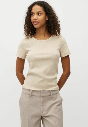 Jeune femme aux cheveux bouclés portant une chemise beige à manches courtes et un pantalon beige à chevrons, debout, les mains dans le dos.