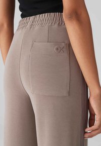 Licht taupe sweatpants met een elastische tailleband, zijrits en een patchzak met een reliëfmerkdetail op de achterkant.