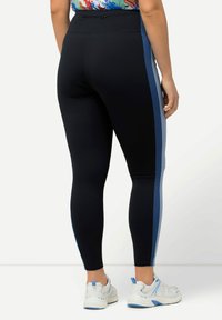 Svarta leggings med blå sidostrikar, slät tyg, tajt design, hög midja, som bärs med vita sneakers. Bakvy visas.