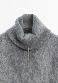 Maglione grigio in cashmere con colletto alto, caratterizzato da una zip frontale e una texture morbida. Etichetta visibile con informazioni sul contenuto e sul marchio.