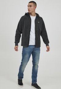 Grau gefütterte Zip-Hoodie mit einer Fronttasche und verstellbarem Kapuze, kombiniert mit hellblauen Jeans und schwarzen Turnschuhen. Einfaches und funktionales Design.