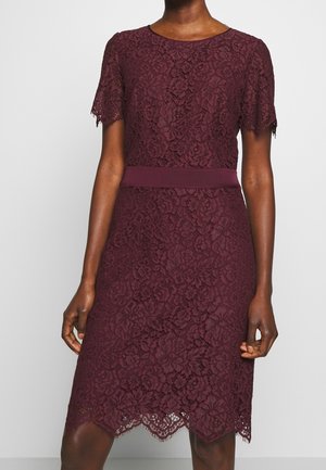 Kobieta w bordowej sukience z koronki o długości do kolan, z krótkimi rękawami, dopasowaną talią i kwiatowym wzorem, stojąca na jednolitym tle.