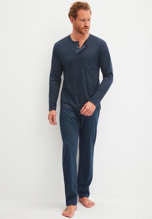 Man met krullend haar, gekleed in een donkerblauw henley-shirt met lange mouwen en donkerblauwe pyjamabroek met patroon, staat op blote voeten tegen een witte muur.