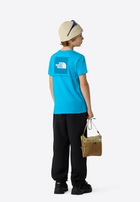 Turquoise t-shirt with a logo on the back, black pants, and a small tan bag. Features a snug fit and cotton texture.