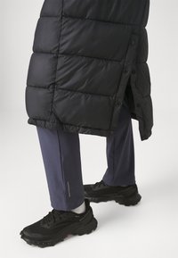Giacca a vento nera con design trapuntato e bottoni, abbinata a pantaloni blu navy con un accento argentato e sneakers nere texturizzate.