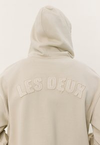 Beige hoodie featuring a front kangaroo pocket and embroidered "LES DEUX" text on the back in textured white stitching.