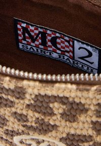 El interior de una bolsa trenzada en color marrón claro, con un forro de tela marrón, presenta una etiqueta rectangular que muestra "MC2 SAINT BARTH" en un diseño a cuadros negro y rojo.