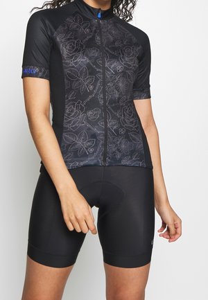 Žena oblečená v černém krátkém cyklistickém dresu s květinovým vzorem a odpovídajících černých polstrovaných cyklistických šortkách stojí před bílým pozadím.