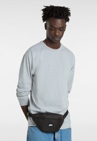 Saku hitam yang dikenakan di pinggang dengan tali yang dapat disesuaikan, menampilkan patch logo. Model mengenakan baju lengan panjang berwarna abu-abu muda.