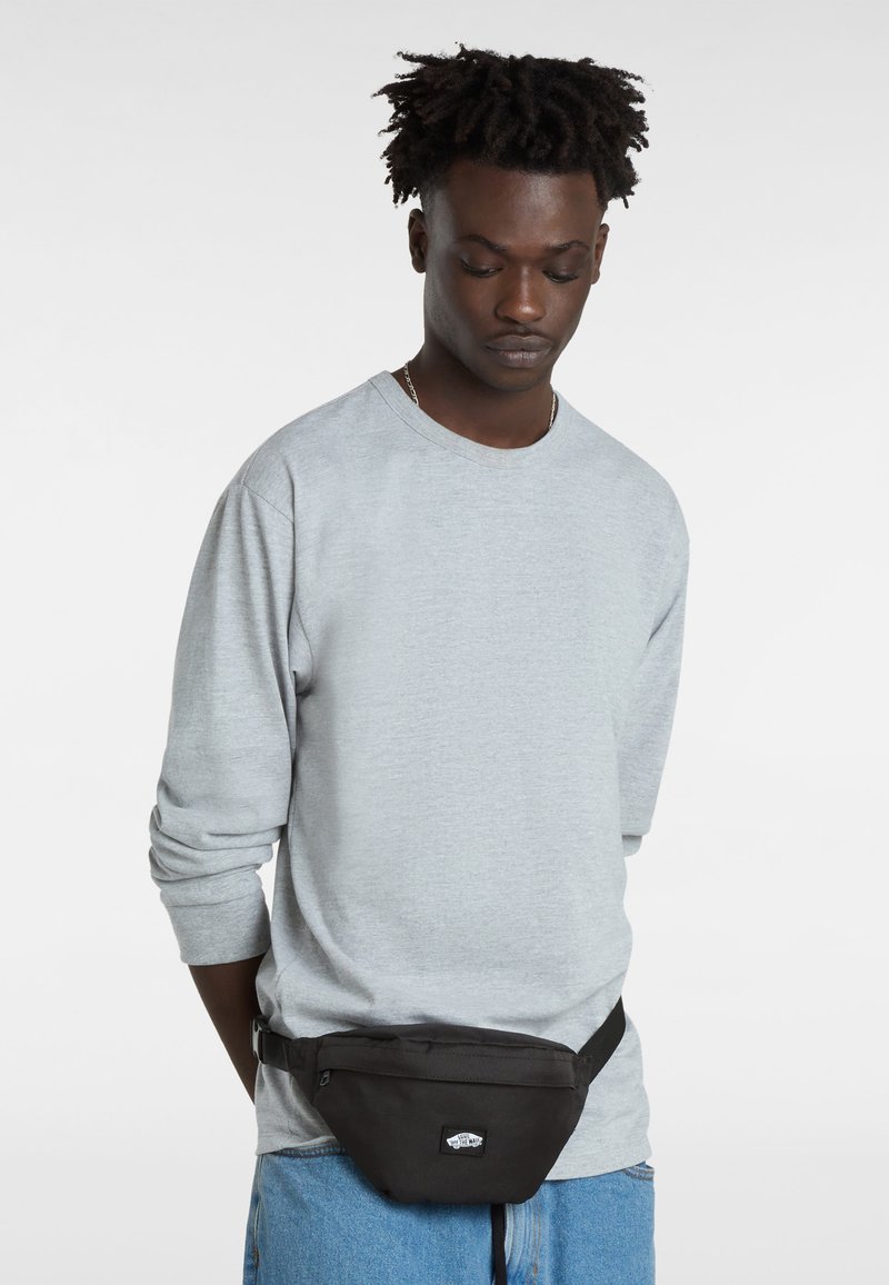 Zwarte stoffen heuptas gedragen om de taille met een verstelbare band, voorzien van een logo patch. Het model draagt een lichtgrijze lange mouwen shirt.