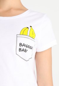 Bílý bavlněný tričko s grafikou kapsy znázorňující dvě žluté banány a textem "BANANA BABY" v černé barvě. Krátké rukávy a kulatý výstřih.