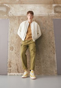 Chaqueta bomber de color claro sobre un suéter naranja, combinada con pantalones cargo oliva y zapatillas blancas con acentos amarillos. De pie contra una pared texturizada.