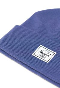 Fialová pletená čepice s žebrovanou texturou. Na přední straně má bílou obdélníkovou nášivku s černým textem. Jednoduchý, skládací design.