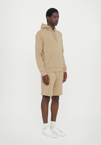 Beige hoodie en shortset, gemaakt van een zachte katoenmix. Bevat een kangoeroezak en een trekkoord in de taille. Gecombineerd met witte sneakers.